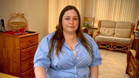 Erin sitting on a chair wearing a blue top. She has long brown hair and a chain around her neck. Behind her is a room filled with wooden furniture. 