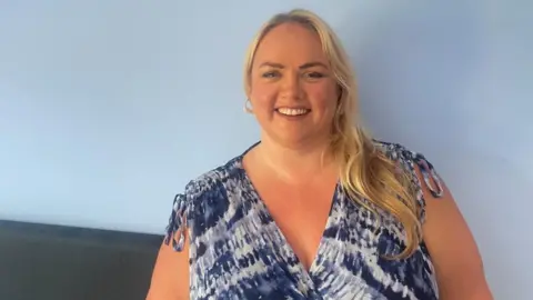 A woman with long blonde hair smiles at the camera. She is wearing a blue and white v-necked top with no sleeves. Behind her is a wall painted light blue.