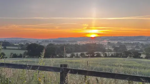 A wooden fence with a green field in front of it and an orange sun rising in the background.