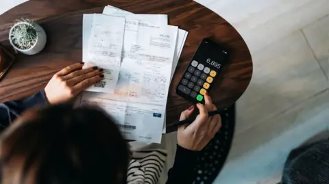 Getty Images A woman looks down at a small stack of bills and a recipt, with a phone calculator near her right hand showing an amount of 6,895.