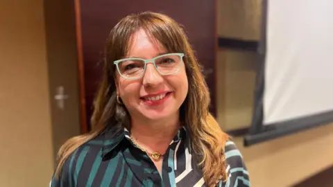 A woman wearing a striped blouse and blue reading glasses smiles at the camera. She is standing indoors. There is a beige wall behind her. 