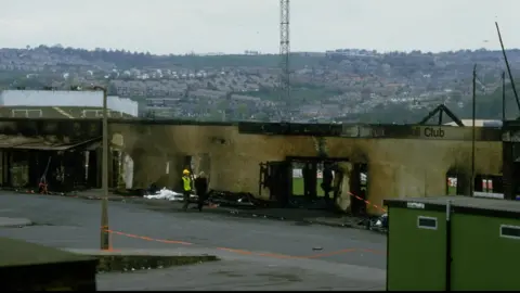 Getty Images A burnt-out stand at Valley Parade's ground in 1985. Two men, one of whom is wearing a hard hat and a luminous jacket, can be seen walking alongside it.
