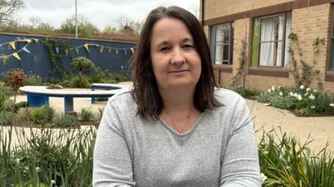 A woman with long brown hair sits in a garden wearing a long-sleeved grey top.