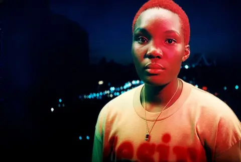 Joshua Gordon Arlo Parks is athed in red light, as she poses against the soft-focus lights of a city at night-time
