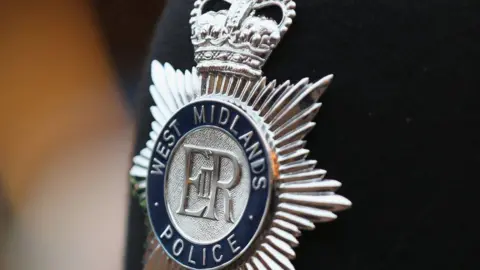 A close-up of a silver police badge on a black jacket, which reads West Midlands Police.
