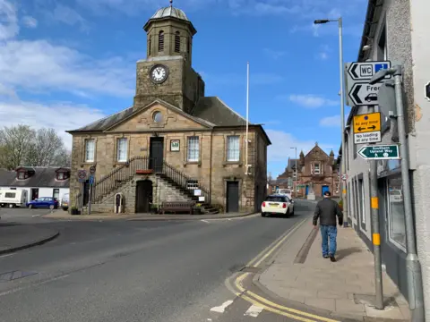 Sanquhar's Tolbooth Museum a large stone building next to a road with a car driving past it. A man is walking along the pavement next to it.