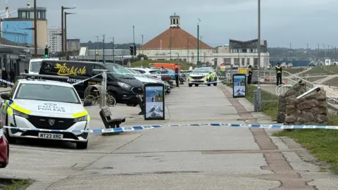 Police tape across the beach-side path in Aberdeen. Two police cars are visible, as well as two uniformed officers