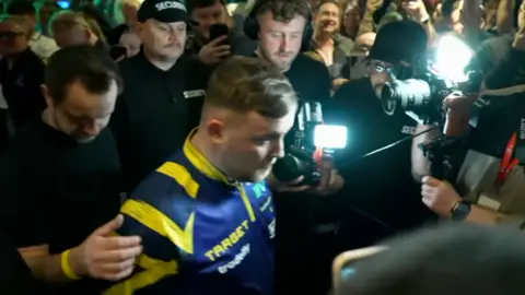 A man in a blue and yellow top walking through a crowded bar with camera lights on him