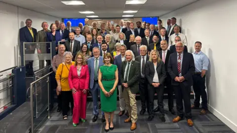 A group of 55 county councillors, dressed smartly, stand in a meeting room, which has white walls and a navy and grey carpet, smiling.
