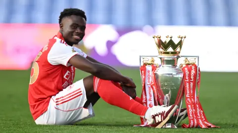 Arsenal's Bukayo Saka sitting next to the Premier League trophy