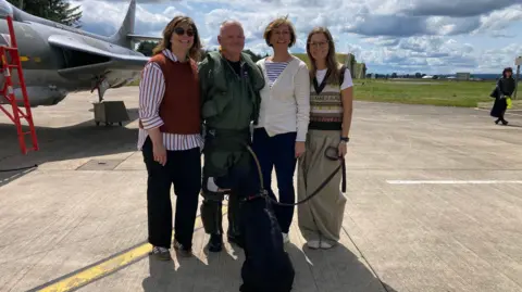 Cathy Killick/BBC A family of one man, three women and a black labradour standing on an airstrip in front of a green fighter jet