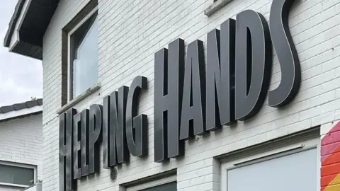 Paintworks NI An exterior wall of the Helping Hands building. It is painted white and the words "Helping Hands" are in capital letters in bold black font