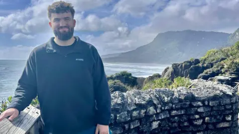 Lloyd Lloyd, who has dark curly hair and facial hair, is wearing a dark jumper and is standing by a stone wall at a coastal viewpoint, with ocean waves, cliffs, and a cloudy blue sky in the background.