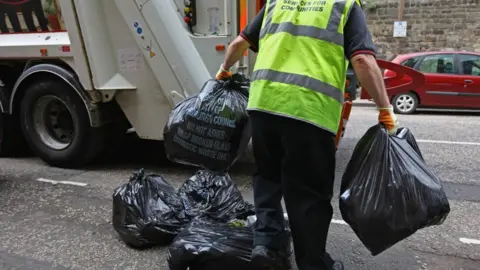 A man in a high vis jacket carries black bin bags to a bin lorry 