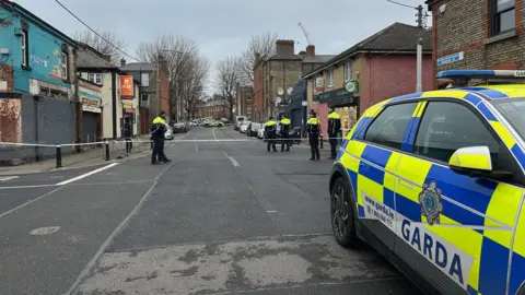 A wide shot of a number of gardaí officers at a police cordon on a street in Stoneybatter, north Dublin. There is a garda car parked on the right hand side of the image.