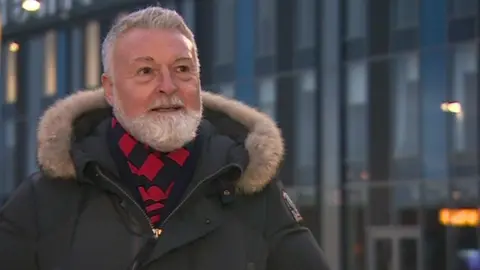 A man, Jason McGill, stands outside the LNER Community Stadium in the evening. He is wearing a coat as well as a red and black striped scarf.