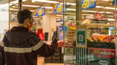 Getty Images A man in a mask scans a vaccine pass QR code outside a shop in Hong Kong