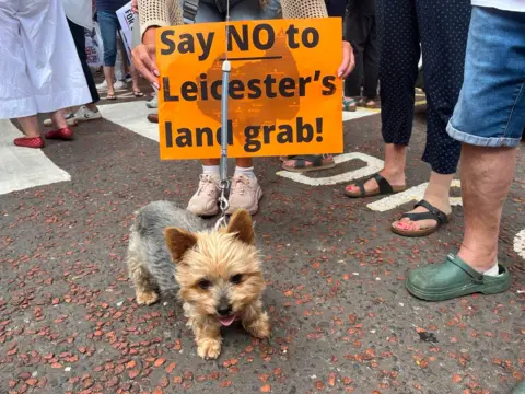A dog with an orange sign saying "say no to Leicester's land grab."