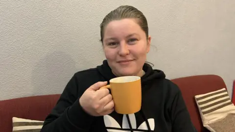 A woman with dark hair scraped back is wearing a black hoodie with a white logo. She is sitting on a red sofa holding a yellow mug.