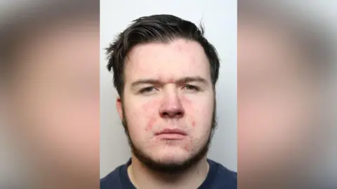 A mug shot of a man with brown hair, a short brown beard and spots on his face. He is wearing a navy t-shirt and is frowning.