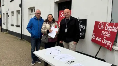 Phil Turner, Justine Norris and Andy Barnes standing outside a white building. They are behind a white table with pens and paper on it. There are black and red placards leaning against the building's external wall with the words: "Gt Burstead needs its own surgery."