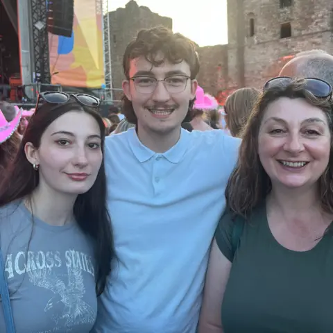 Family A young woman with brown hair in a grey shirt stands next to a young man with glasses and a light blue shirt, who is also next to a woman with brown hair and a green shirt. The two women, who are a mother and daughter, both have sunglasses pushed up onto their heads