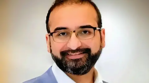 Family A man with short dark hair, beard and moustache and wearing glasses, in a portrait photo against a light background. A white collar and blue jacket are visible at the base of the photo.