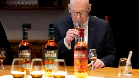John Swinney is a bald man wearing glasses and a dark suit. He holds up a glass of whisky to his nose and appears to sniff it. A couple of drams and bottles of Loch Lomond whisky sit on a table in front of him.