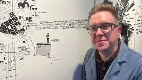 Andy Boreham, wearing a blue jacket and round glasses, standing next to the 'story wall' in Bristol Beacon, which has illustrations on it showing previous performances.