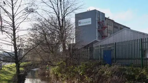 The outside of Futamura Chemical UK Ltd factory site. It is a factory with silver ridge panelling and a large building attached to it. There is high green iron fencing surrounding the site, with a small river to the left of it with some trees and shrubbery.