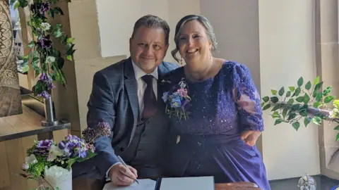 David and Allison Friday on their wedding day. They are standing smiling broadly while signing official papers. David has short dark hair and is wearing a suit. Allison is wearing a purple dress and has an up-do. Her arms are amputated below the elbow. 