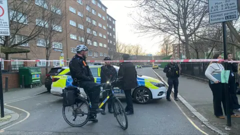 BBC police car and officer on a bike next to Red Police Cordon with police activity on the road