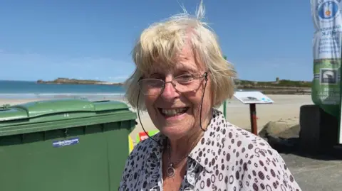 Hilda, a woman in seventies to eighties, is smiling. She has glasses on, shorts grey/blonde hair and is wearing a white and brown spotty shirt. In the background there is a green wheelie bin and a view of the bay.