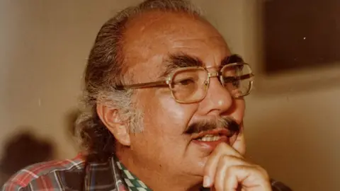 A close-up colour half-profile shot from the right of a man in his mid-60s, looking away to the right of frame with his chin in his hand. He has grey hair which is still dark in places, and a moustache which is still mostly dark, and dark bushy eyebrows. He is wearing glasses, a watch with a silver strap, a red, white and grey tartan-patterned shirt, and a white and green neckerchief.