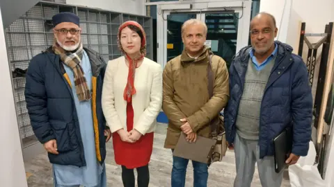 Three men and two women standing in a line, smiling at the camera, with a set of glass doors behind them.