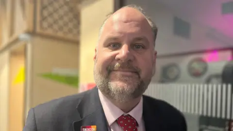 Jim Robbins smiles at the camera - he is wearing a grey suit jacket, pink shirt and red tie.
