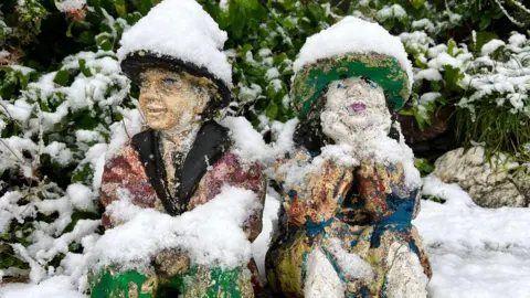 Snow has covered two garden ornaments of a boy and girl. The ornaments are both wearing a hat which has been covered in snow. Snow has blanketed a bush behind them.