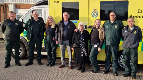 East of England Ambulance Service Eight people smiling, standing in line, facing the camera, with an ambulance behind them. A man and woman with grey hair are in the middle, and are flanked by two women and four men, who are all in ambulance service uniform. 