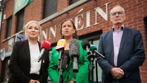 Liam McBurney/PA Wire Sinn Fein President Mary Lou McDonald TD (centre), with First Minister Michelle O'Neill (left) and Gerry Kelly MLA (right), speaking to the media outside the Sinn Fein offices in Belfast in September 2025