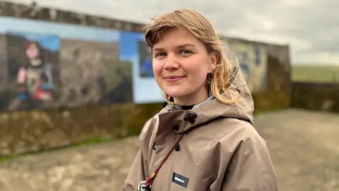 BBC A young woman with blonde hair, smiling at the camera. She is wearing a beige rain jacket.