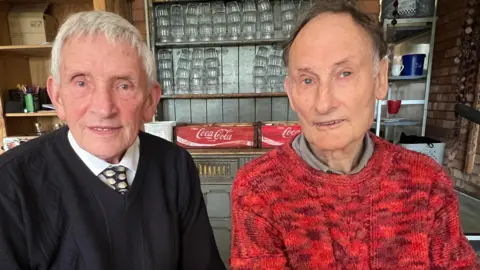 Roger and his friend Gerald looking at the camera smiling and wearing jumpers and shirts but Gerald is wearing a tie