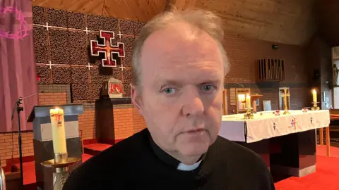 Fr Gormely has thin, greying hair, and wears the black short and white collar of a priest. Behind him is an altar, lectern and assortment of holy candles. 