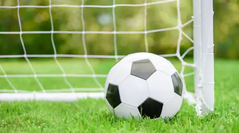 Getty Images A generic photo of a black and white football near a football post and white net, with bushes and grass blurred in the background.