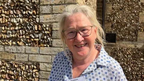 Vikki Irwin/BBC Barbara Buckley wearing a light coloured top smiling at the camera with a stone building behind her.