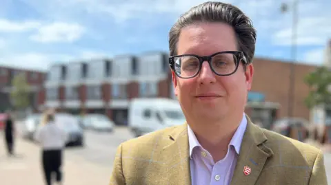 Andrew Schrader wears a checked light brown suit jacket and a purple shirt. He has black-framed glasses. He looks at the camera. A road is behind him.