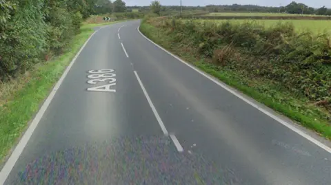 The image shows a quiet rural road curving gently to the left through countryside.
