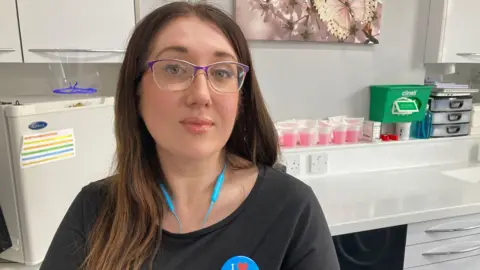 A woman with brown long hair and glasses with a purple edge is looking at the camera. Behind her is a fridge, cups with pink liquid and a green medical box.