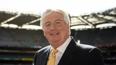 A man with short grey hair stands with his right arm resting on a large silver bowl trophy, with red and navy ribbons around the trophy's handles. Behind him is a large empty stadium with a large green pitch in the centre. The man is wearing a black suit, white shirt and yellow tie.