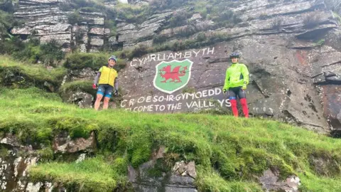 Tom Gregory Tom and a man standing high above the Rhondda in front of a sign saying Cymru am byth, Welsh forever and Croeso ir Gymoedd, welcome to the Valleys. The picture is taken from a distance. Both are wearing cycling outfits.