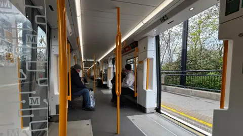 Passengers on board of a new Tyne and Wear Metro carriage. Yellow grab poles are space out across the centre of the train, which has linear London tube-style seating. There are also poles next to the doors. 
